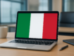 A laptop displaying the Italian flag on the screen, placed on a wooden desk with a coffee cup and notebook nearby.