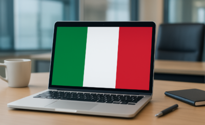 A laptop displaying the Italian flag on the screen, placed on a wooden desk with a coffee cup and notebook nearby.